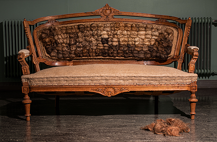 Marja Kanervon installaatio Uudelleenverhoilu, jossa on vanha sohva verhoiltuna hiuksilla sekä kasa hiuksia sen edessä.