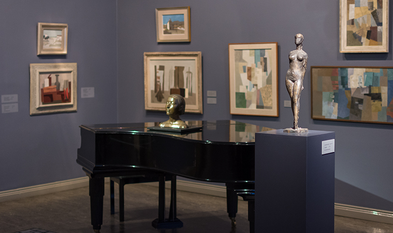 Kuvassa on näkymä Joensuun taidemuseon Resetti-huoneesta, jonka sinisenharmaalle seinälle on ripustettu kubistisia, vaalean, sinisen ja ruskeiden sävyjä sisältäviä maalauksia. Niiden edessä on musta piano, jonka päällä on pronssinen naisen päätä esittävä veistos. Kuvan etualalla on jalustalla alastonta naista esittävä pieni pronssiveistos.